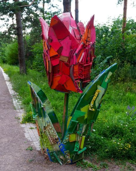 Eine große Tulpen-Skulptur aus bunten Altmetallstücken steht am Rande eines Weges in einem grünen Park.