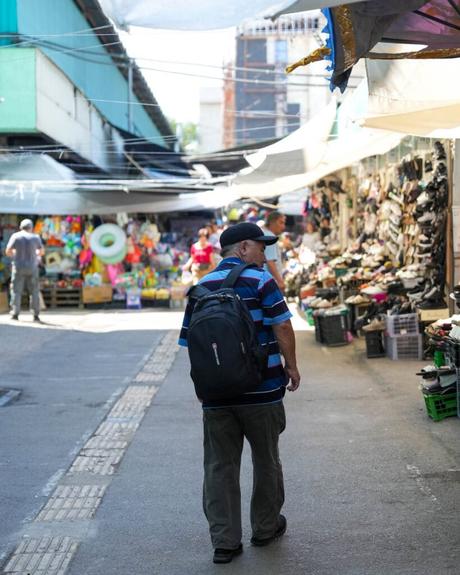 Ein Mann mit einem Rucksack geht durch einen Markt im Freien, an dem Stände mit Kleidung, Schuhen und Accessoires aufgereiht sind.
