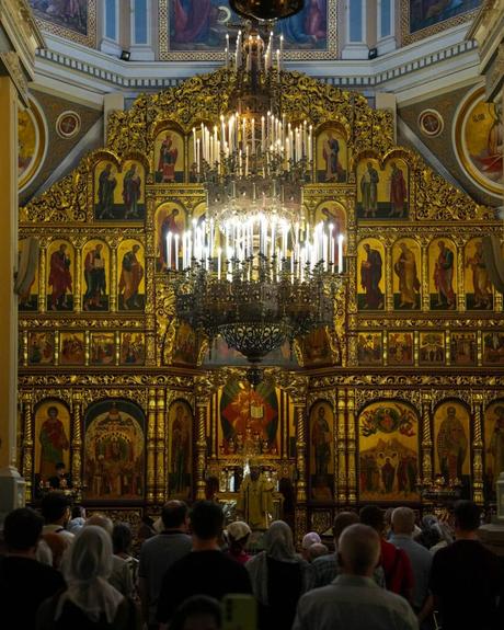 Die Menschen nehmen an einem Gottesdienst in einer prunkvollen Kirche mit goldenen Ikonen und einem großen Kronleuchter an der Decke teil.