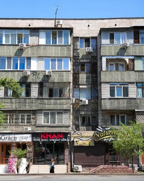 Fünfstöckiges Wohnhaus mit Geschäften im Erdgeschoss; eine Person geht an den Bäumen davor vorbei.