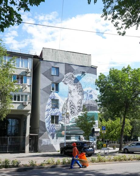 Ein Wandgemälde, das eine große Katze an einem Gebäude zeigt; unten auf der sonnigen Straße schiebt eine Person in orangefarbener Kleidung einen Wagen.