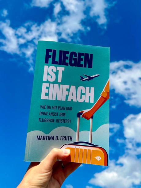 Vor einem blauen Himmel mit Wolken im Hintergrund hält eine Hand ein deutsches Buch über das Fliegen in der Hand.