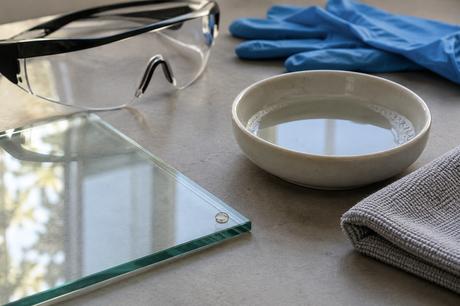 Eine transparente Platte mit kleinem Tropfen liegt neben einer Wasserschale, Schutzbrille, Handschuhen und Mikrofasertuch.
