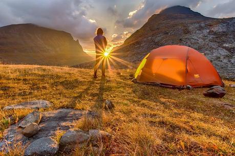 Ein Mann steht neben seinem Zelt während einer Wandertour und bewundert den Sonnenuntergang zwischen den Bergen