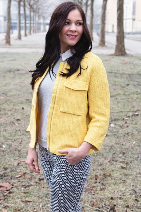 Kleidermädchen präsentiert einen frühlingshaften Look mit gelber Jacke und Musterhosen. Dazu trägt sie flache Slipper und einen grauen Pullover von Vila. 