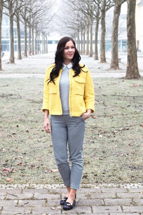 Kleidermädchen präsentiert einen frühlingshaften Look mit gelber Jacke und Musterhosen. Dazu trägt sie flache Slipper und einen grauen Pullover von Vila. 