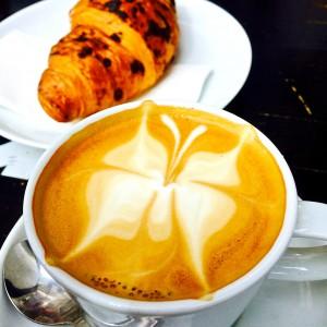 Cappuccino mit Schmetterlingmuster im Milchschaum, Schokocroissant im Hintergrund